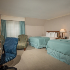 Two queen beds with teal blankets with white pillows near brown armchair and desk with blue desk chair in Ogunquit, ME