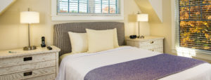 Bed with white pillows, white linens and blue blanket near two bedside tables with lamp and window in Ogunquit, ME.