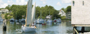 A Group of Different Sail Boats with people on The Ocean with Houses in Background in Ogunquit, ME