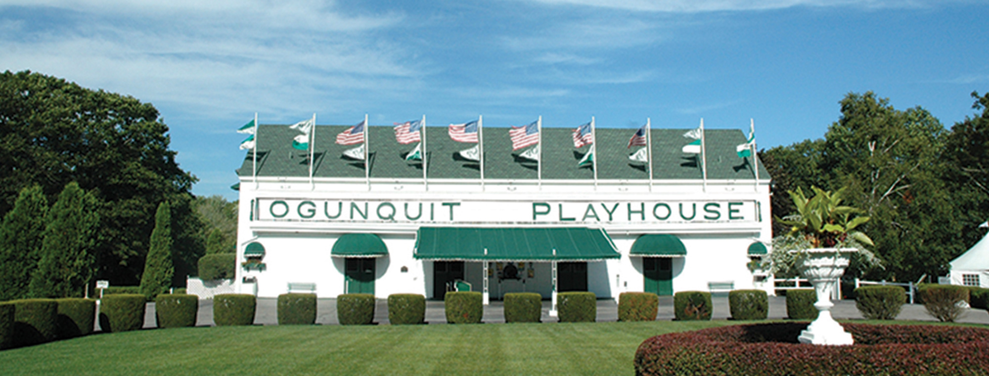 Entrance to Ogunquit Playhouse with flags hanging about building entrance in front of mowed lawn in Ogunquit, ME