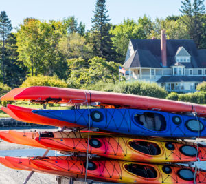 A Rack of Different Colored Kayaks on Rack Located on Beach with House in Background